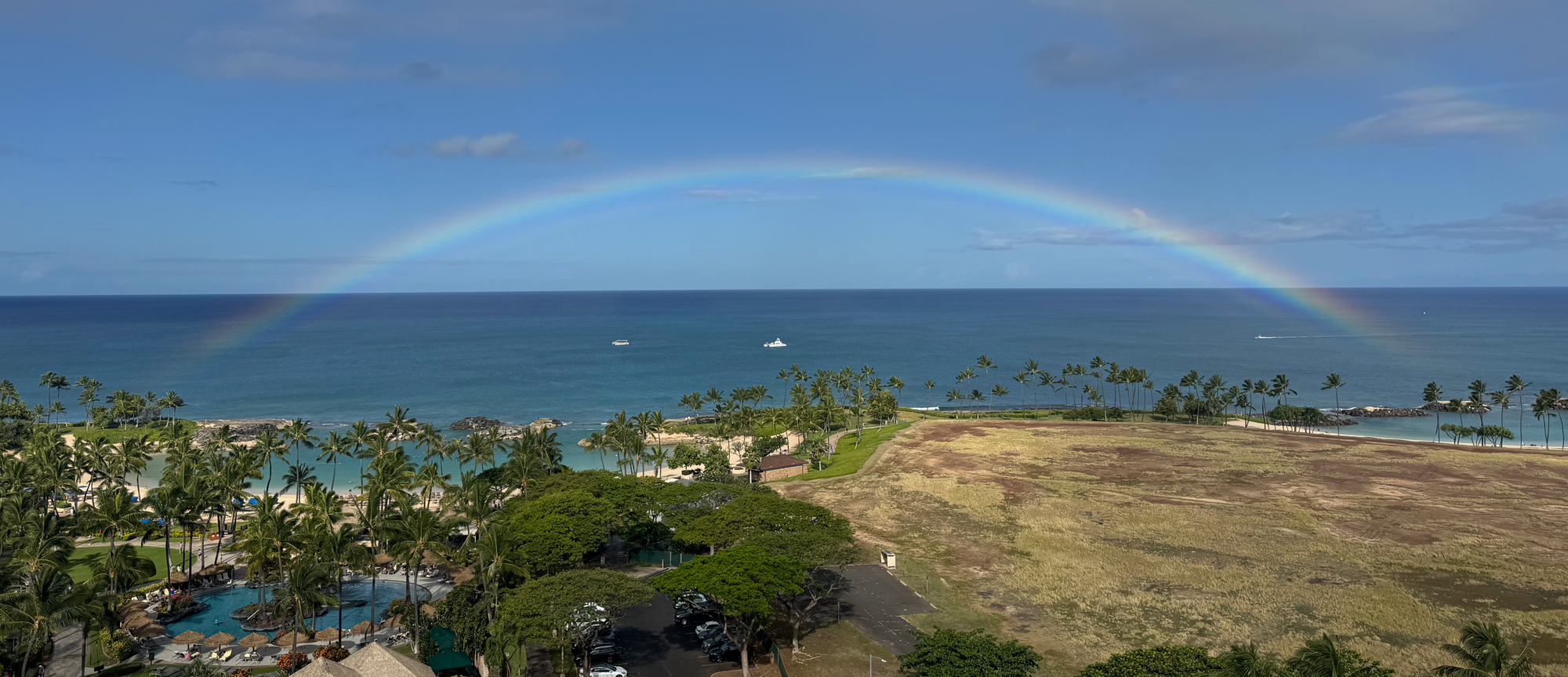 Photograph of a full arc rainbow off the coast of Oahu. The pale rainbow colors contrast with the bright green of palm trees to the left and the brown-green of empty grassland on the right, with a bright blue ocean and sky as the background.