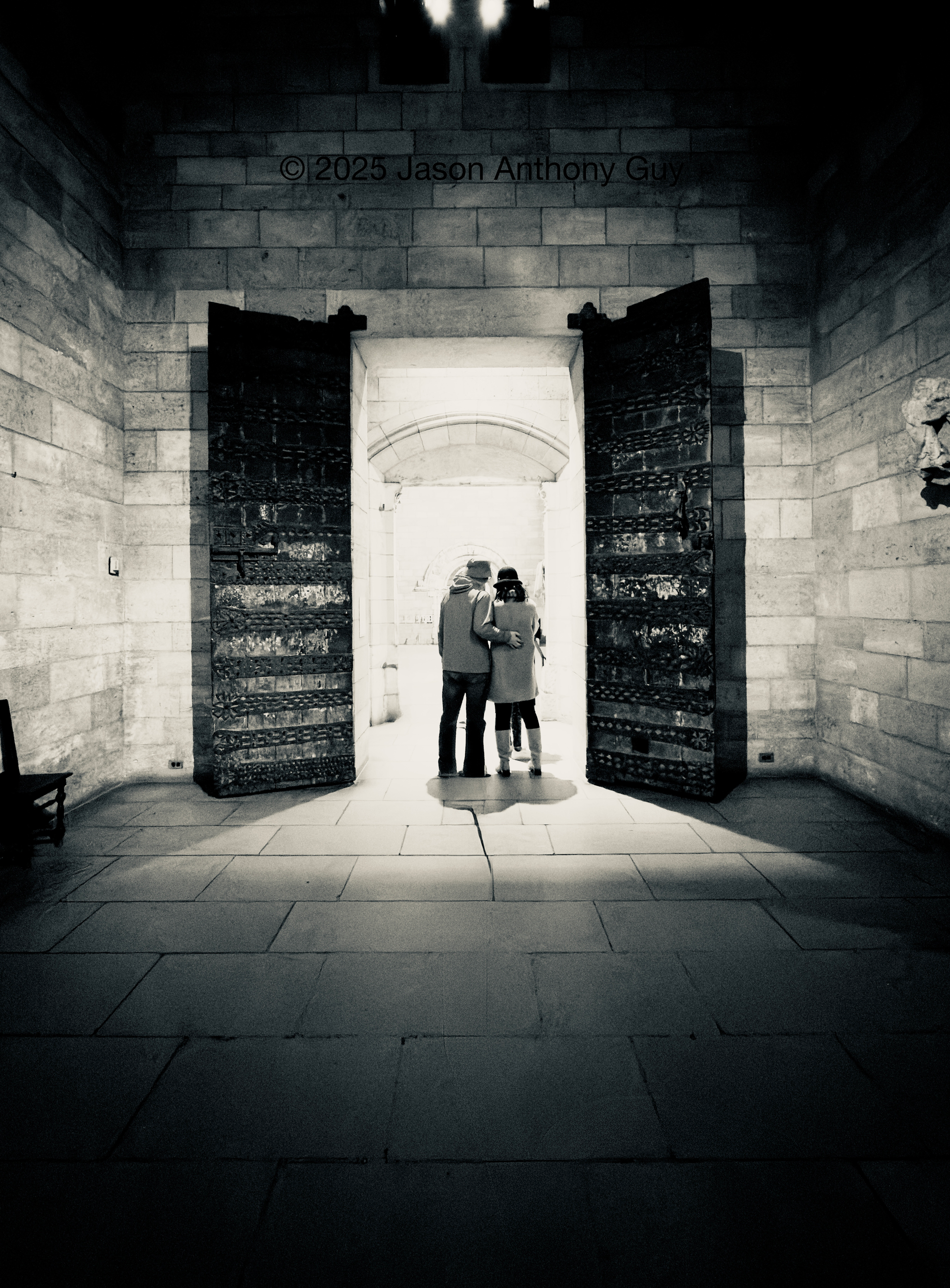 Black and white photograph of a couple, their backs to us, standing in a doorway with ornate doors, under a bright pool of light  The doors are dark wood or metal, which contrasts with the light colored bricks of the building. The light fades off quickly, leaving the foreground in shadow.
