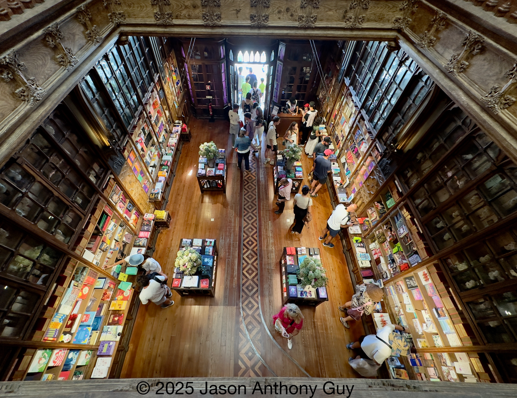 Photography from above looking down to the lower level through ornate wood railings. The brown wood melds into brown shelves, which morphs into colorful and brightly lit books. People mill about examining books.