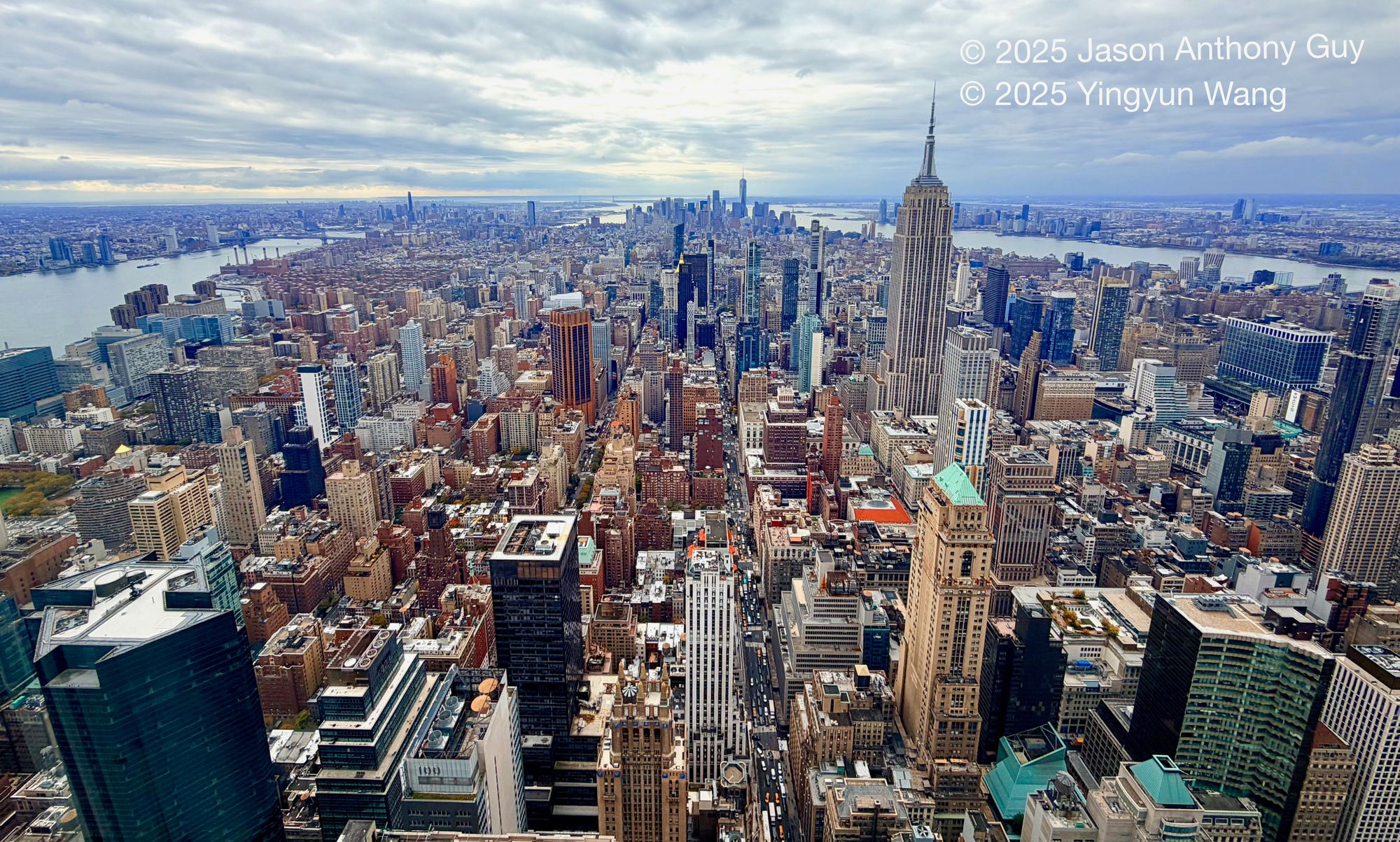 Photograph of New York City, from midtown looking south, with the East and Hudson rivers flanking the island. The grey-blue skies and river contrast with the dusky browns and tans of the buildings. The image stretches out to a vanishing point in the distance.