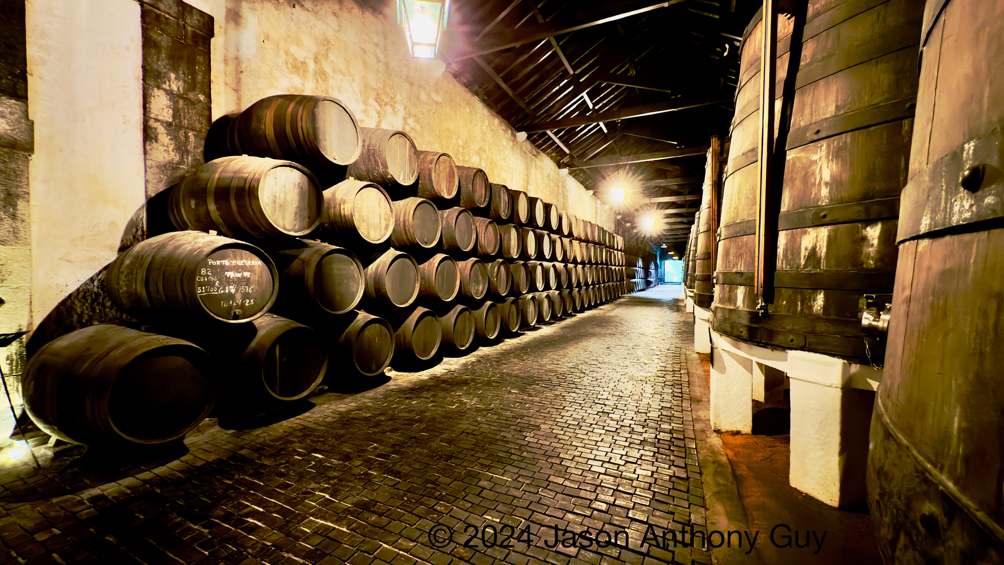 Photograph of dozens of brown wooden barrels stacked on the left, large wooden vats to the right, with a long brick path leading out a door between them. The image is sepia-toned, evoking age and sentimentality.