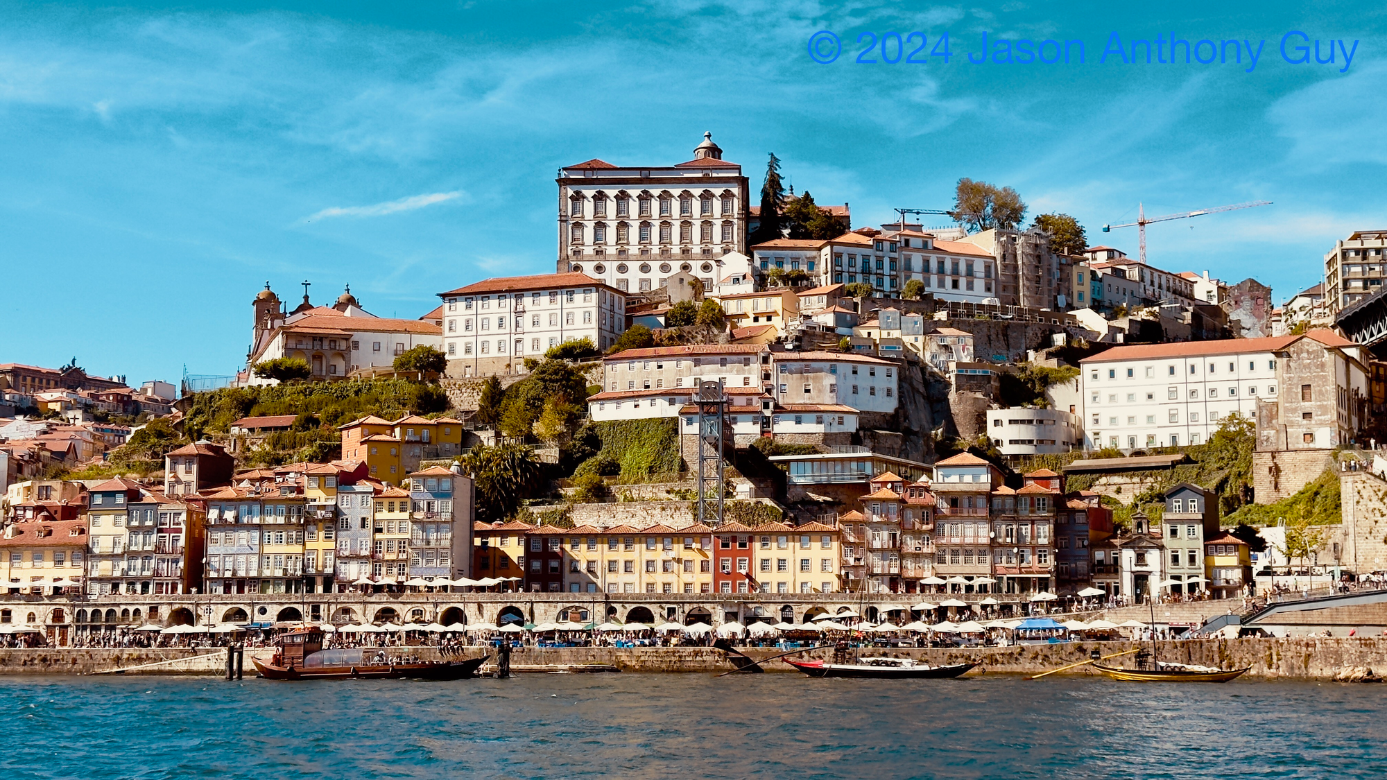 Photograph of several colorful buildings on a hill overlooking a river. The building are in yellows, reds, and creams, which contrasts with the dark blue of the river and the lighter blue of the sky.