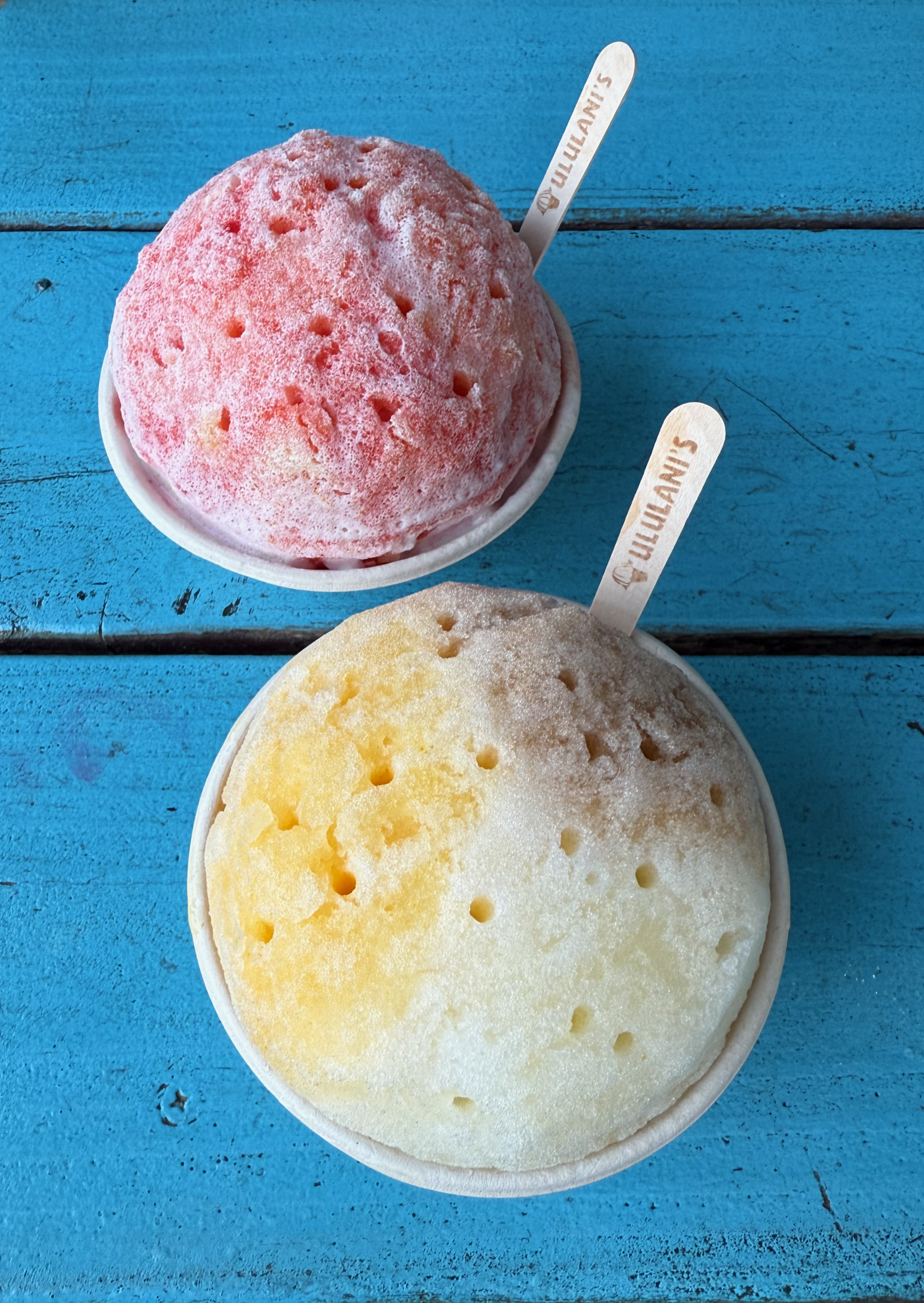 Photograph from above of two cups of shave ice, one red, one yellow and white, sitting on a blue wooden table. Both have a wooden spoon with “Ululani’s” printed on them.
