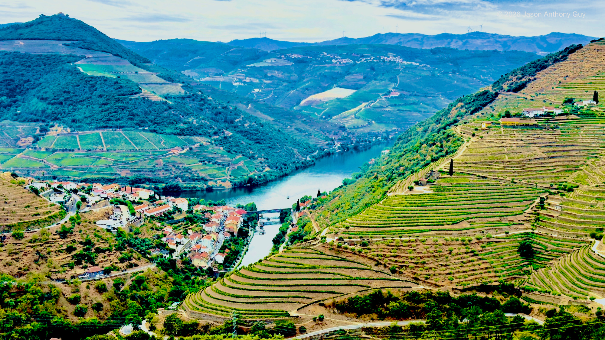 A high, panoramic view of a river winding through a steep valley of terraced hillsides. The slopes on both sides are carved into bands of vineyards and farmland, shifting from bight greens on one side to blue-green in the distance.
